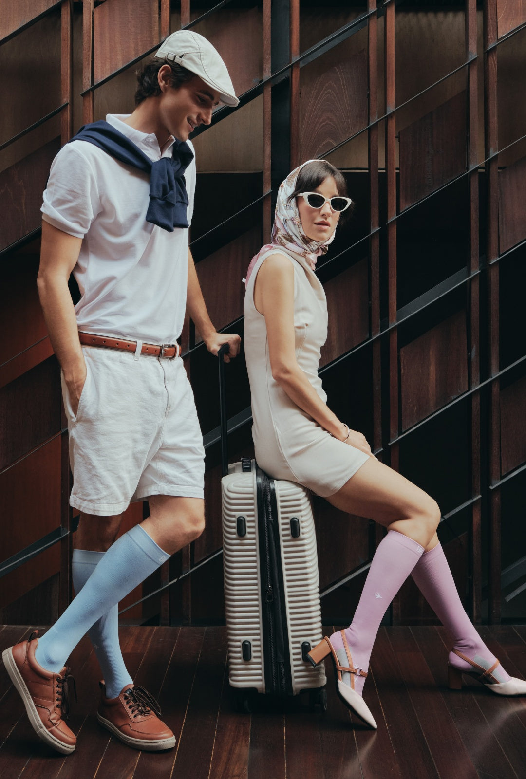 A man and a woman pose with a striped hard-shell suitcase in a modern interior with dark wood paneling. The man wears BIOSx Flight Compression Socks in Sky Blue, and brown shoes. The woman wears the same compression socks in Luxe Lilac and high heels.