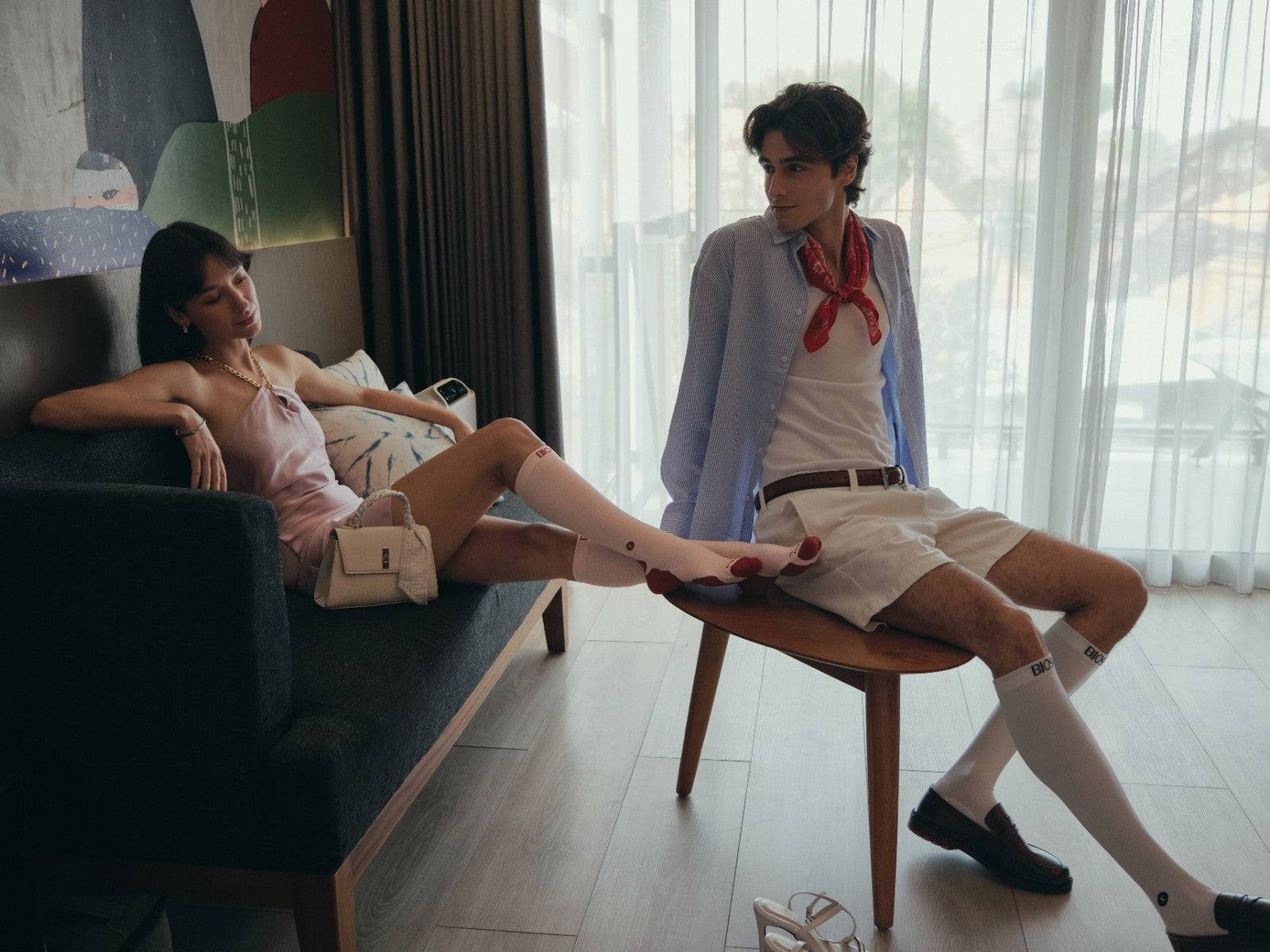 A man and a woman in a stylishly dressed, relaxed indoor setting. The woman is reclining on a dark sofa wearing a light pink dress, pnk thigh-high BIOSx Compression socks, and a small beige handbag. The man wears white BIOSx Compression Socks.